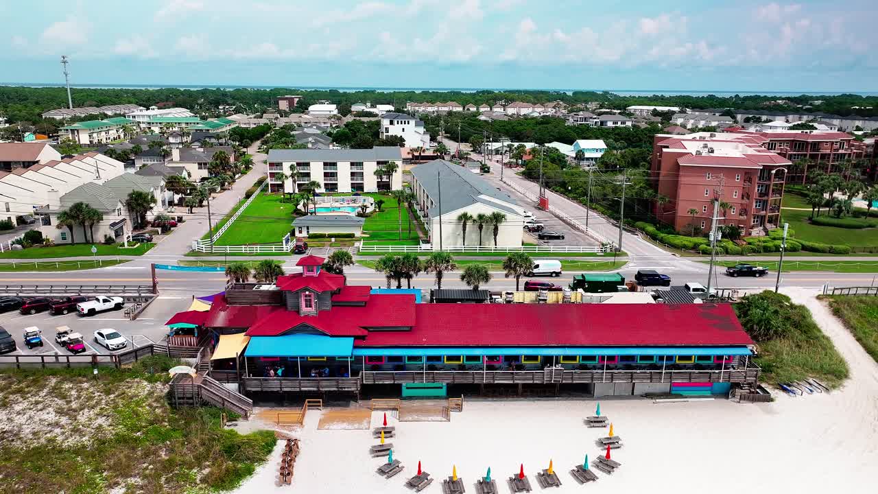 Pompano Joe's restaurant and parking lot trucking right aerial drone shot with a view of old 98, white sand, emerald green water and lots of umbrellas and beach chairs in Destin Florida