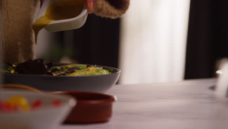 close up of woman at home in kitchen preparing healthy vegetarian or vegan meal pouring dressing onto bowl of salad leaves