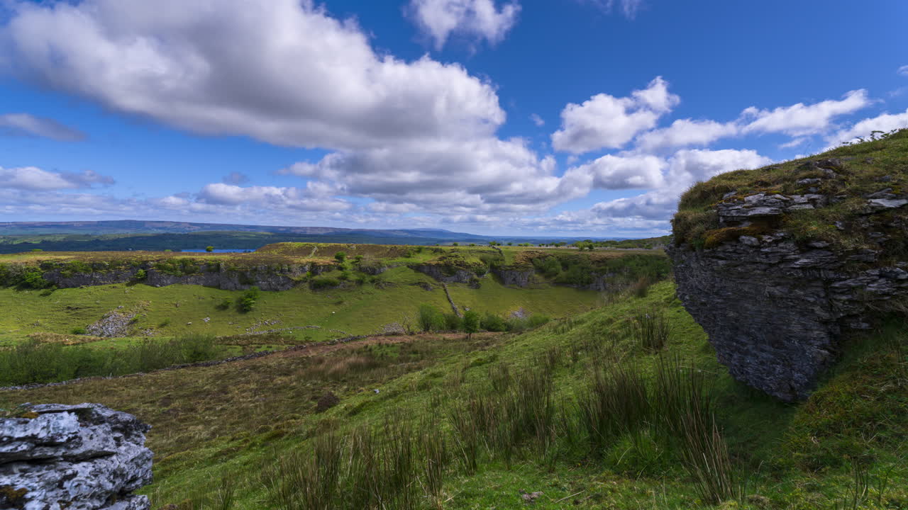Panorama-timelapse af et landligt landskab med klippeblokke og træer i forgrunden og en sø i horisonten p\[unkt] en solrig dag med skyer, set fra Carrowkeel i County Sligo i Irland.