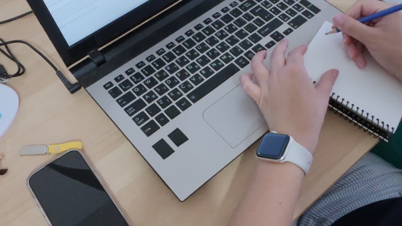 Close-up of hands multitasking, writing in notebook while using laptop.
