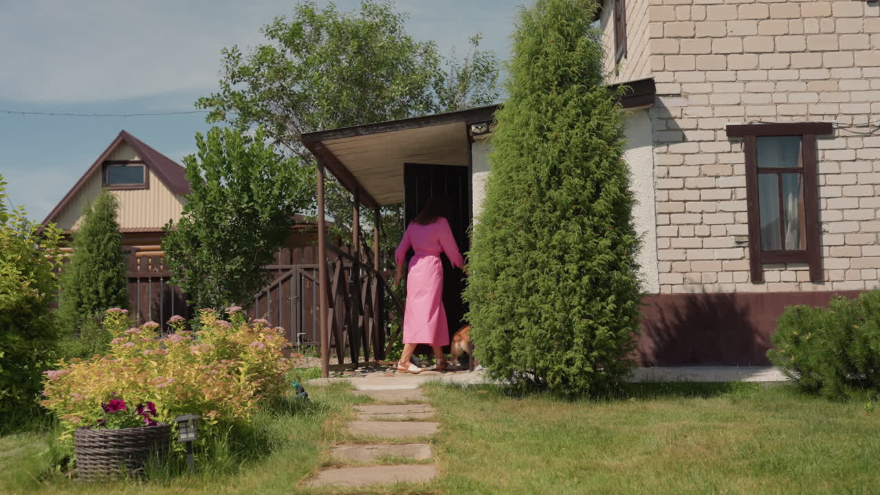 Caucasian Owner Steps Out With Corgi From Shaded Porch, Floral Pots And Trimmed Hedge Around, Smiling Child Trailing Behind Sunlit Morning, Casual Suburban Routine And Pet Companionship