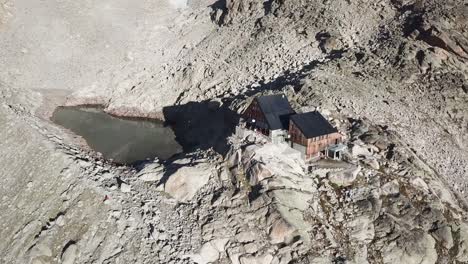 Wooden-hut-next-to-a-lake-for-hikers-and-mountaineers-next-to-rocky-summits-in-the-swiss-alps,-drone-shot