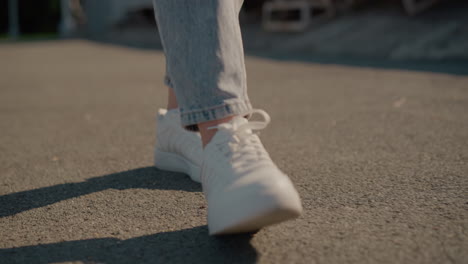 close-up of person walking on tarred road wearing white sneakers and jeans, sunlight casting soft shadows on ground, capturing movement, casual style