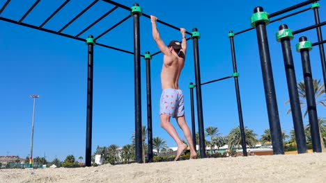 A young male wearing shorts, a baseball hat and sunglasses doing pull-ups on bars at an outdoor gym on the beach