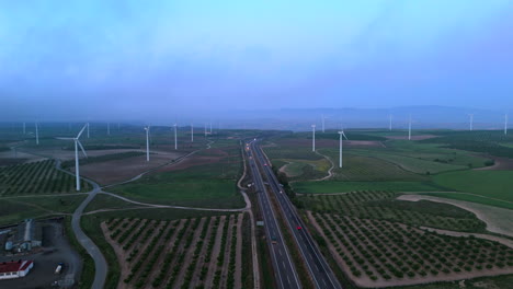 drone flies backward over highway crossing agricultural fields and wind turbines at dawn, with soft light and cloudy sky in a wide rural landscape