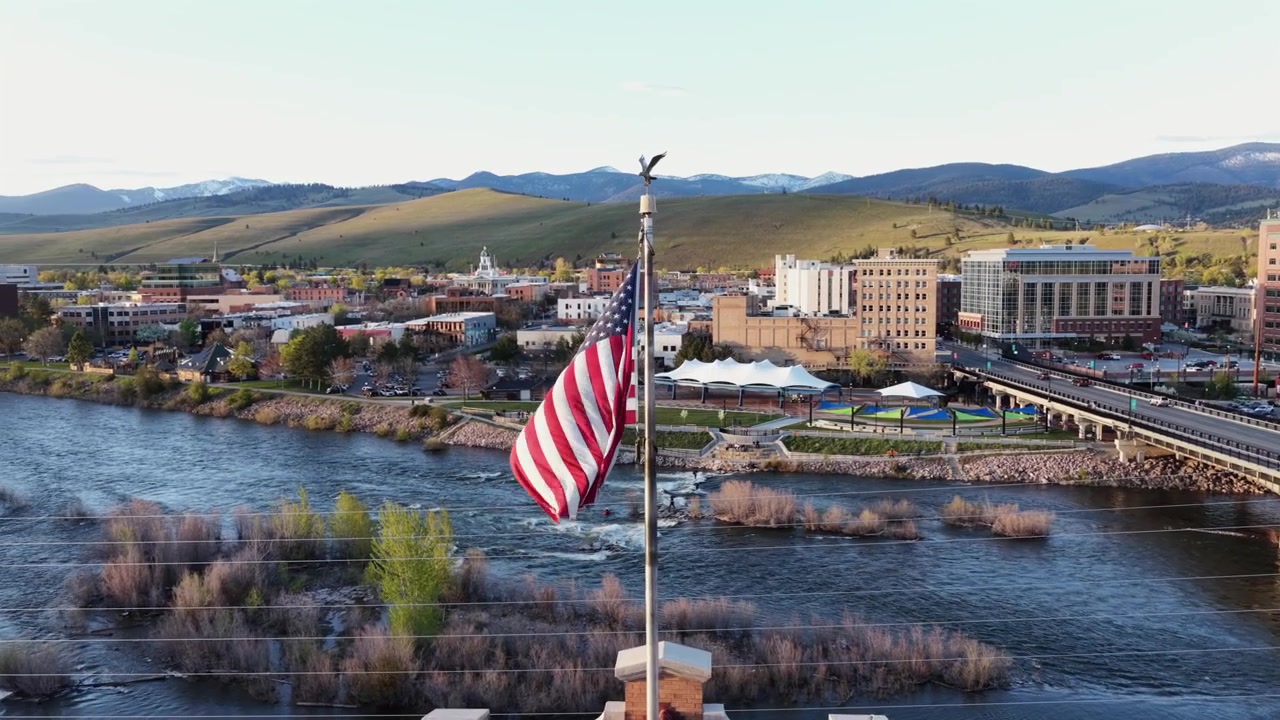 Missoulan keskusta Amerikan lipun, Clark Fork -joen, sillan, joenrantapuiston, tiilirakennusten ja kumpuilevien kukkuloiden kera, lumihuippuisen vuorijonon alla kirkkaan aurinkoisen taivaan alla. Montana, USA. Kiertävä ilmakuva.