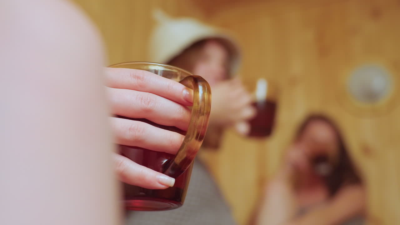 Close up of delicate hand holding glass mug filled with herbal tea in sauna while in blurred background two women wrapped in towels make cheerful toast