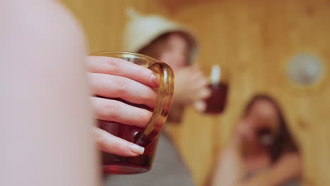 close up of delicate hand holding glass mug filled with herbal tea in sauna while in blurred background two women wrapped in towels make cheerful toast