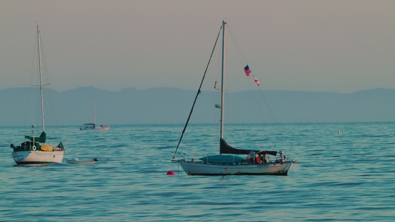 Tranquil sailboat drifts near Santa Barbara, proudly flying American, Canadian, and Red Cross flags under soft evening coastal light.