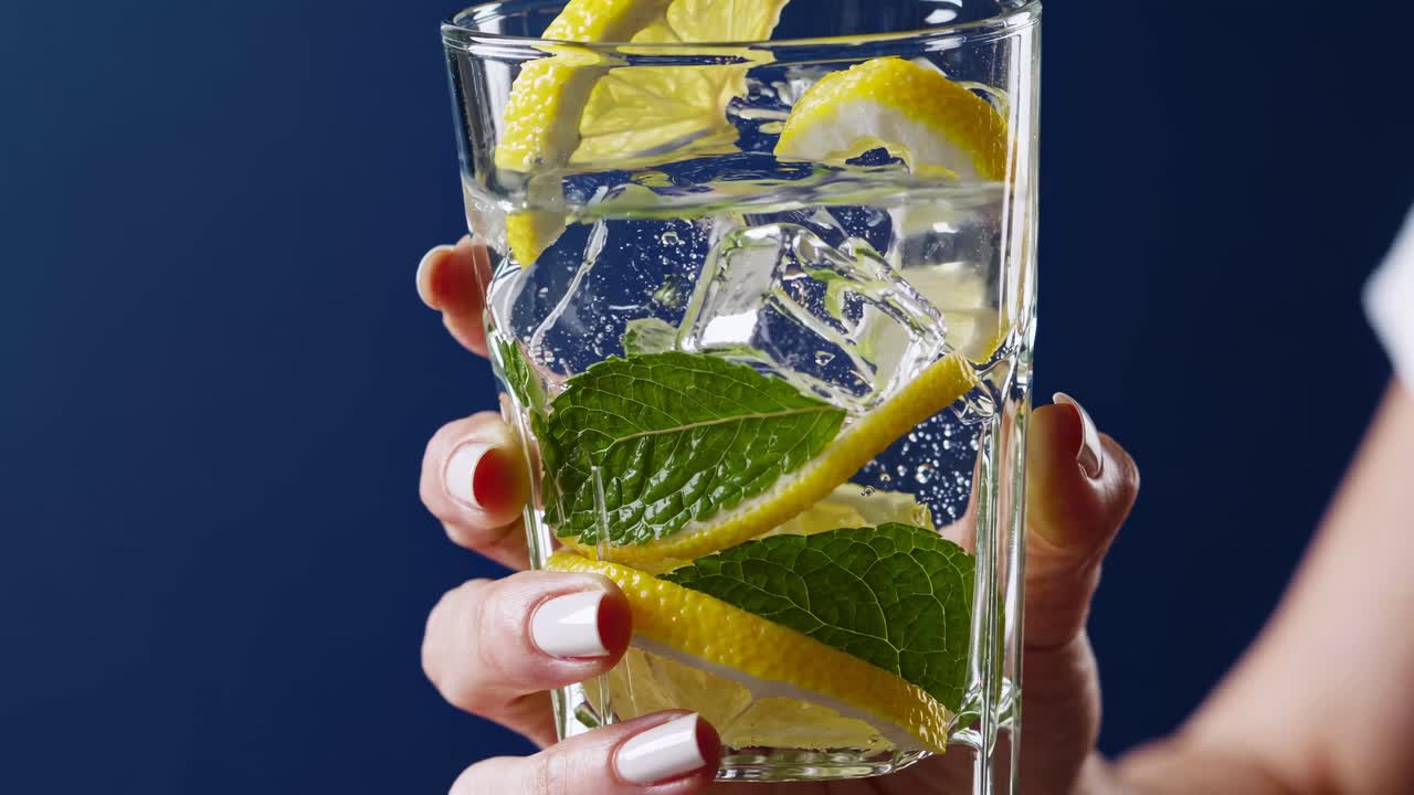 Close-up angle of a hand holding a glass with lemon slices, mint, and ice against a blue background
