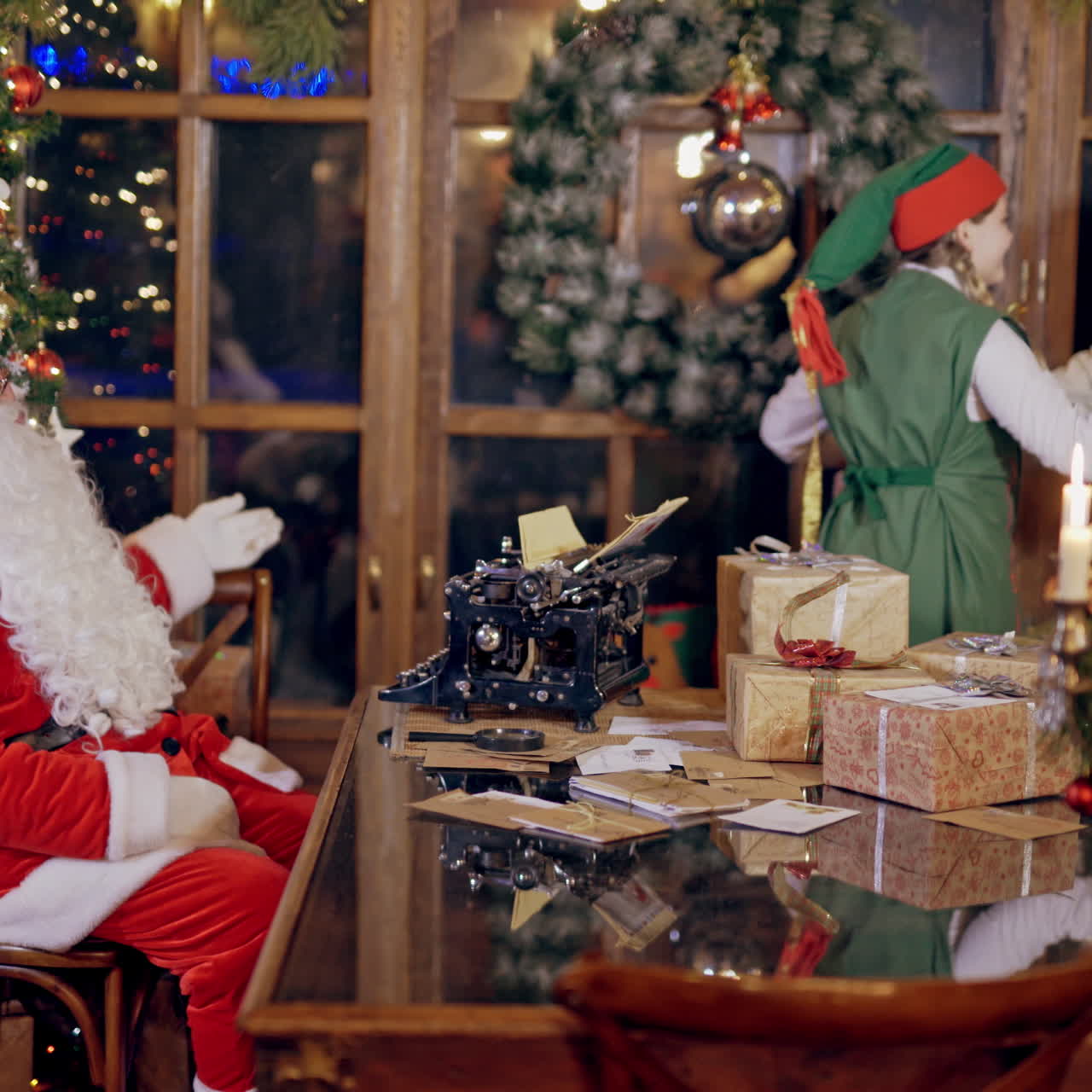 Santa sitting at the table and two elves dance together. Happy elves in fairy costumes dancing while Santa Claus is watching at them on christmas background indoors.