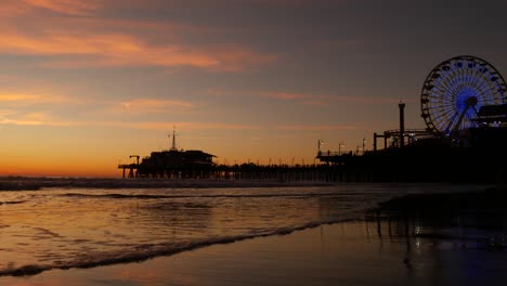 olas del crepúsculo contra la clásica rueda de la ferris iluminada, parque de atracciones en el muelle en santa mónica pacific ocean beach resort. símbolo icónico del verano de california brillando al anochecer, los ángeles, ca ee.uu.