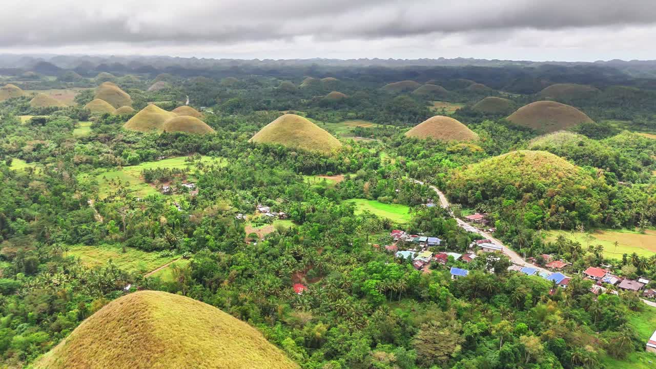Stunning aerial pull out shot reveals the iconic Chocolate Hills of Bohol, Philippines, scattered across lush tropical forest and villages under dramatic overcast skies on a humid day