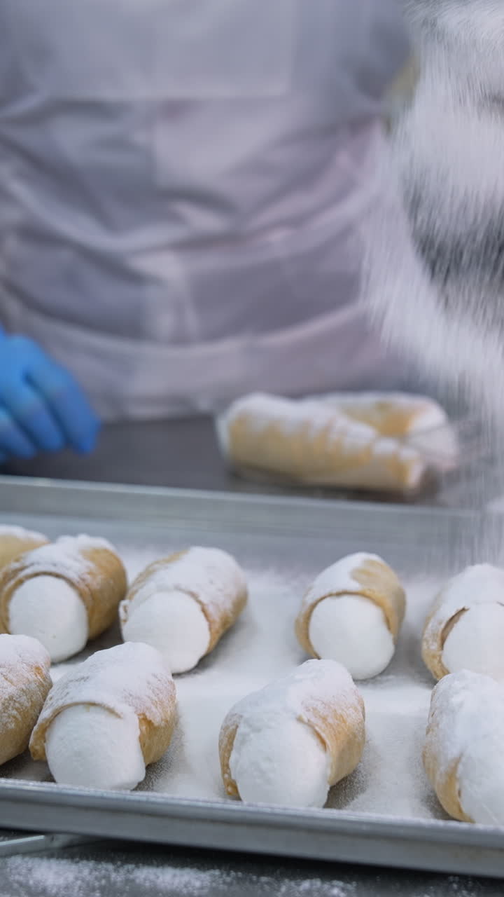 Savory cannoli sweets laid on the metal tray for decoration. Worker covers desserts with sugar powder from a sieve. Close up. Vertical video