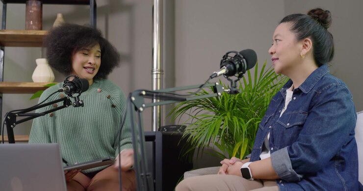 African American and Asian women co-hosting in studio, smiling, holding tablet and speaking on mic