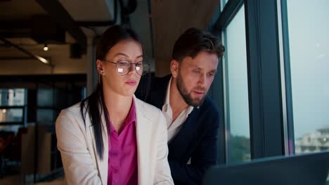 A-brunette-girl-in-a-white-jacket-and-pink-shirt-with-round-glasses-sits-with-a-guy-in-a-blue-jacket-and-works-on-a-laptop-near-a-panoramic-window-in-the-office