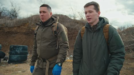 two confident military men in green uniforms and with backpacks on their backs walk along a combat training ground with a barricade of car tires and communicate in the steppe