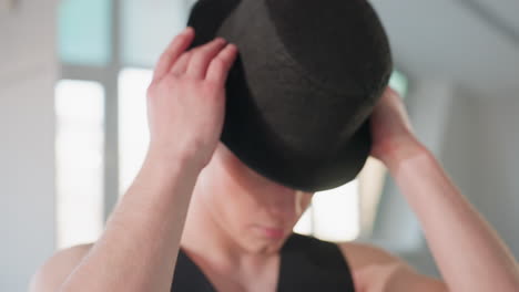close up of young man retrieving black cap from chair and putting it on while standing in sunlit loft studio, mirror reflecting gesture and room details, focus on hands and motion