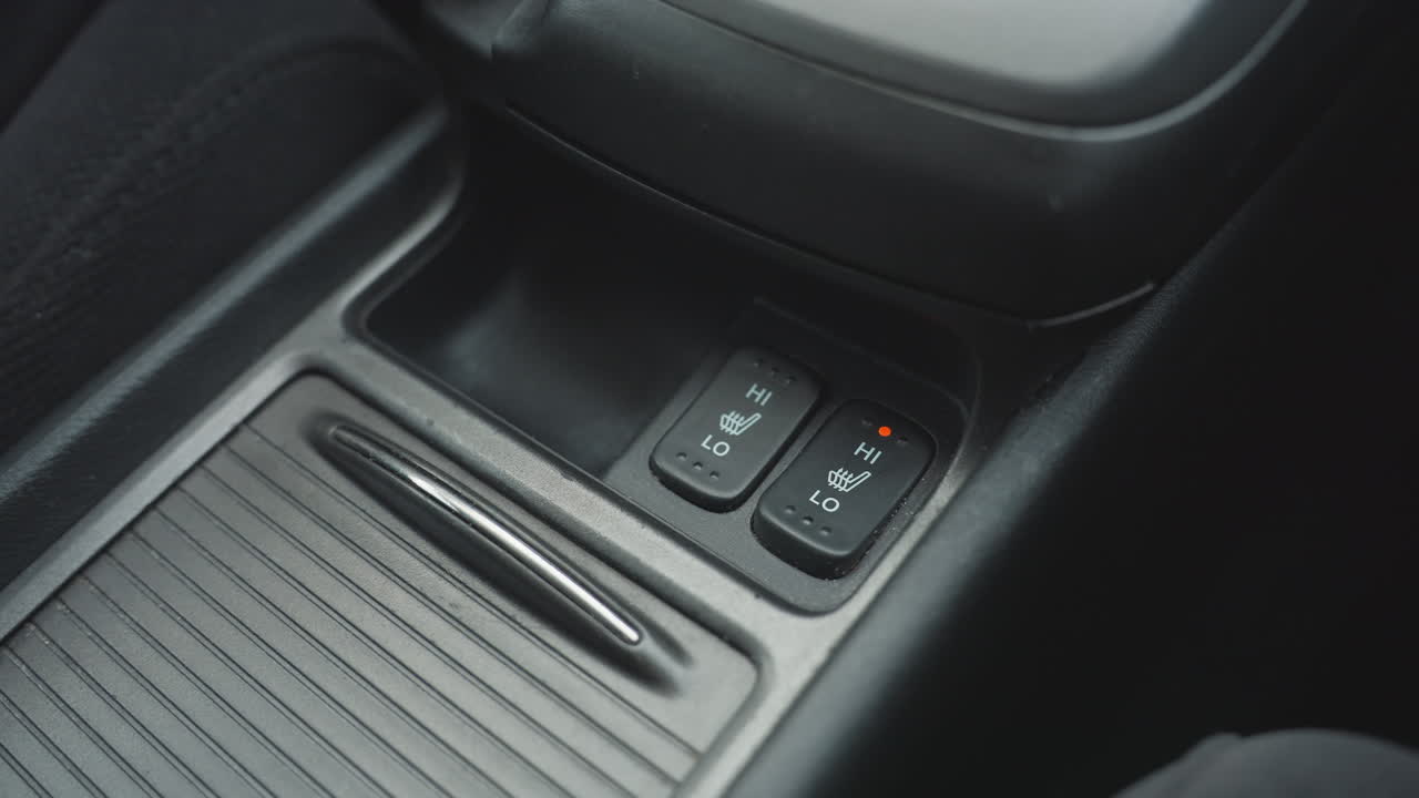 Close up of person switching off one seat heater button and switching on adjacent seat heating control inside car center console showing finger press and visual indicator light activation