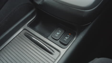 close up of person switching off one seat heater button and switching on adjacent seat heating control inside car center console showing finger press and visual indicator light activation