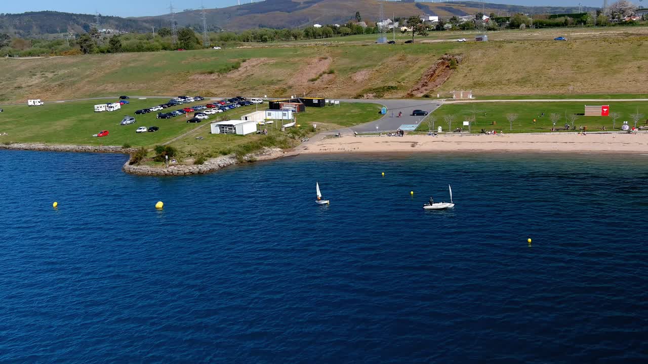 gente practicando en la escuela de vela del lago con playa, aparcamiento y aerogeneradores en las montañas al fondo