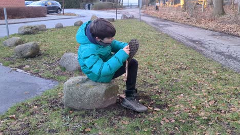 Boy-is-Sitting-on-a-Stone-Cleaning-His-Dirty-Boots-With-a-Stick