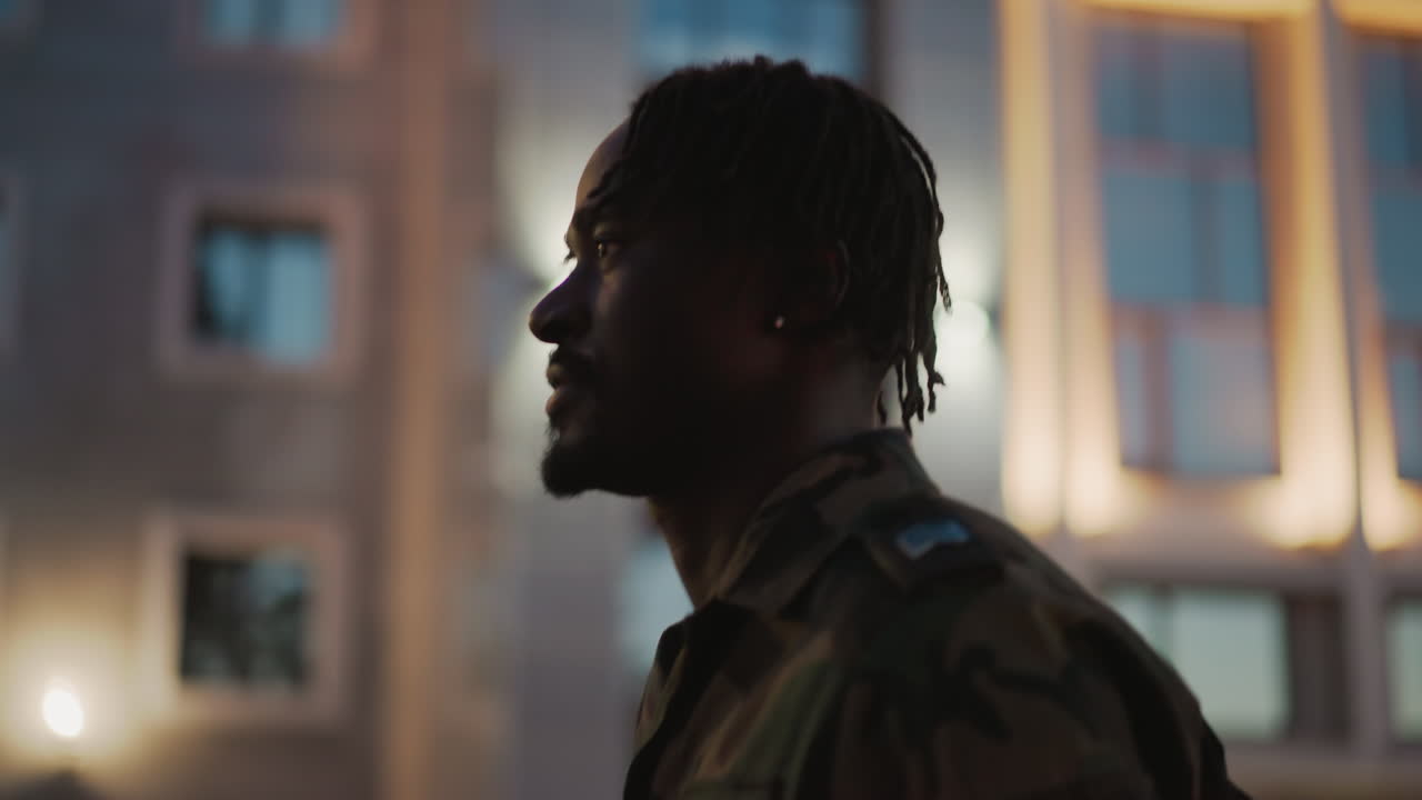 Soldier Journeys Home, Camouflaged Figure Marches In Evening, Soldier In Camouflage Walking Past Glowing City Windows, Former Soldier In Camouflage Strides Past Illuminated Buildings At Dusk