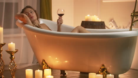 Woman-lying-in-bubble-bath-with-the-light-from-the-candles-to-relax-and-drink-wine.-Romantic-relaxing-aromatherapy-atmosphere.-Bathroom-with-candles.