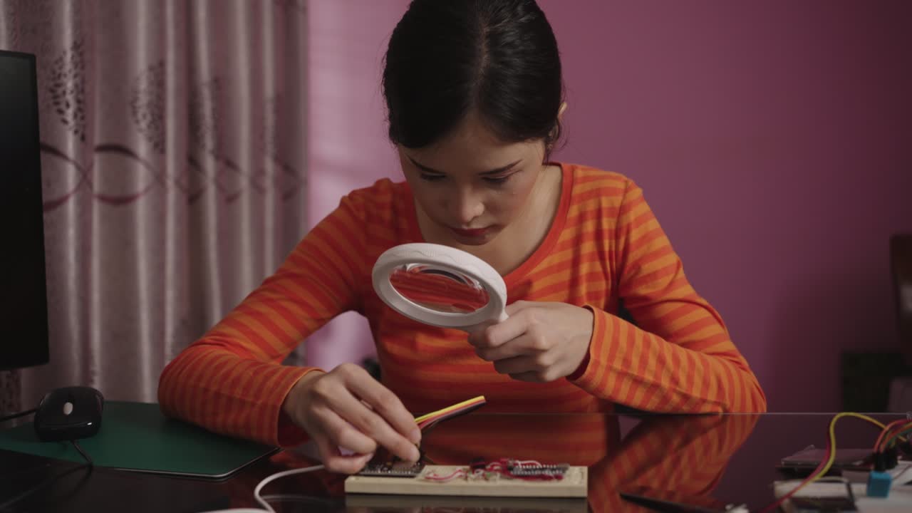 Asian woman uses a magnifying glass to plugging a pin on microcontroller board. Internet of things.