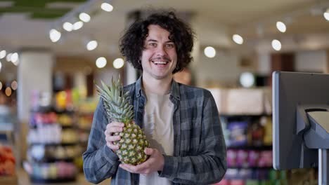 Portrait-of-a-happy-brunette-guy-with-curly-hair-in-a-plaid-shirt-who-holds-a-pineapple-in-his-hands-and-smiles-broadly-in-a-supermarket