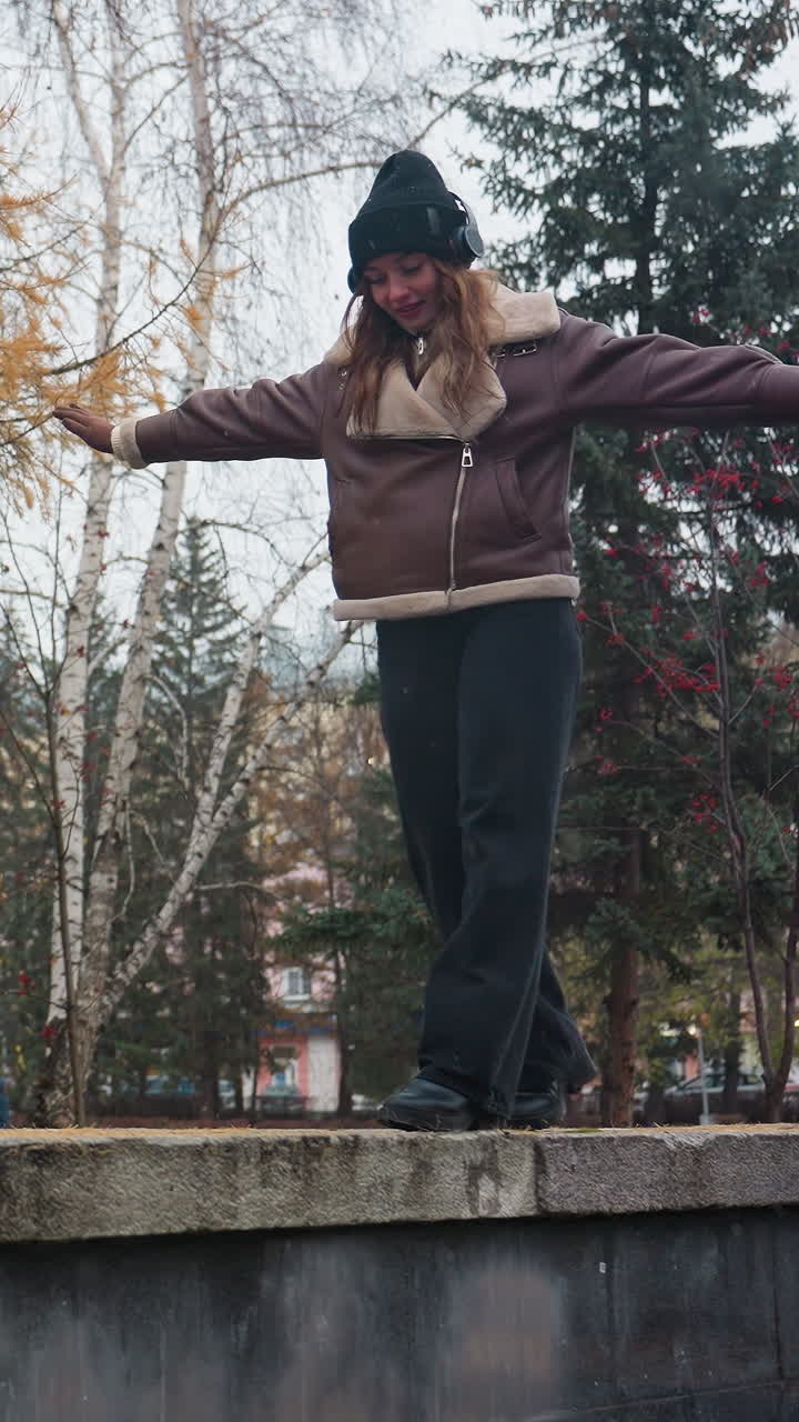 Dreamy lady wearing black knit cap brown shearling jacket black trousers balancing gracefully with arms outstretched while walking on wide stone surface in quiet autumn park filled with colorful trees