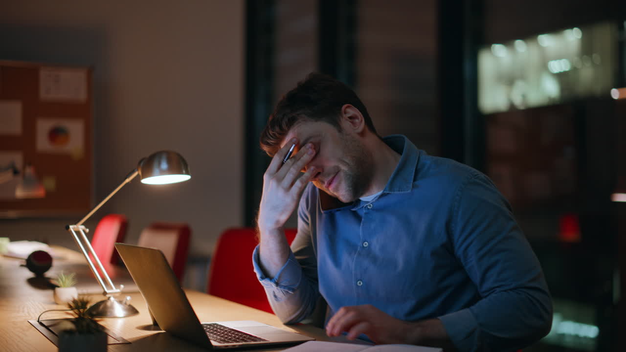 Uomo sovraccarico di lavoro che parla al telefono e legge appunti al computer portatile in un ufficio buio - primo piano