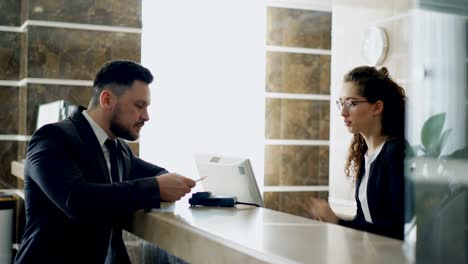 businessman guest talking with receptionist girl standing at desk in hotel reception and paying bill with credit card via contactless payment technology . business, travel and people concept