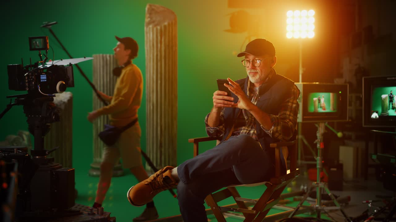 유명한 감독이 휴식시간에 의자에 앉아 스마트폰을 사용하고, 고급 장비를 장착한 스튜디오에서 전문 제작진이 고예산 영화를 촬영하고, 카메라를 어보고 있다.