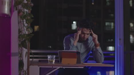 Late-night-overworked-and-stressed-man-doing-head-massage-sitting-in-front-of-his-tablet-on-the-balcony