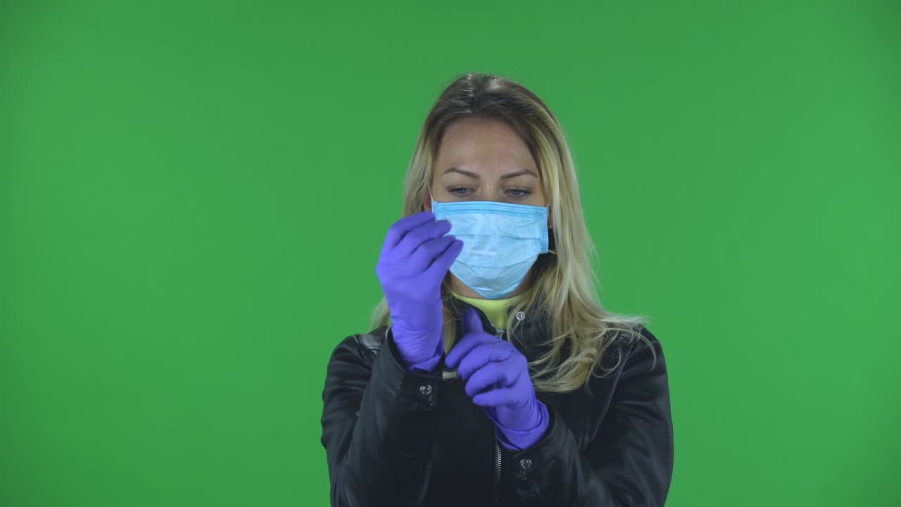 retrato de una hermosa mujer joven con máscara médica está mirando a la cámara y se pone guantes de goma. rubia con cabello suelto en una chaqueta negra y vaqueros en una pantalla verde en el estudio. protección de la salud concepto de virus corona