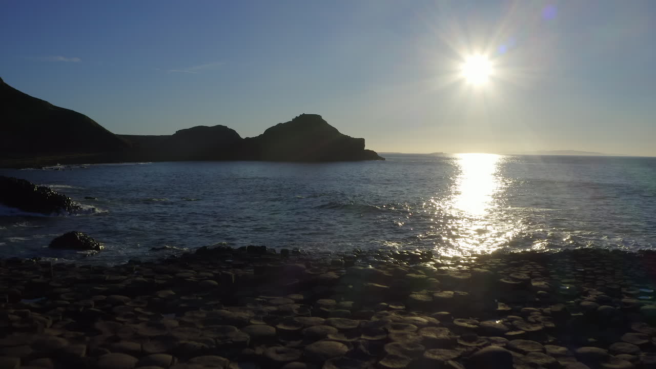 Sunset illuminates the basalt steps at Giant's Causeway as the drone glides forward, framing the sun