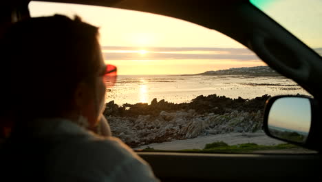 Frau-Im-Auto-Zeigt-Auf-Etwas-Im-Meer,-Während-Die-Sonne-über-Dem-Ozean-Untergeht