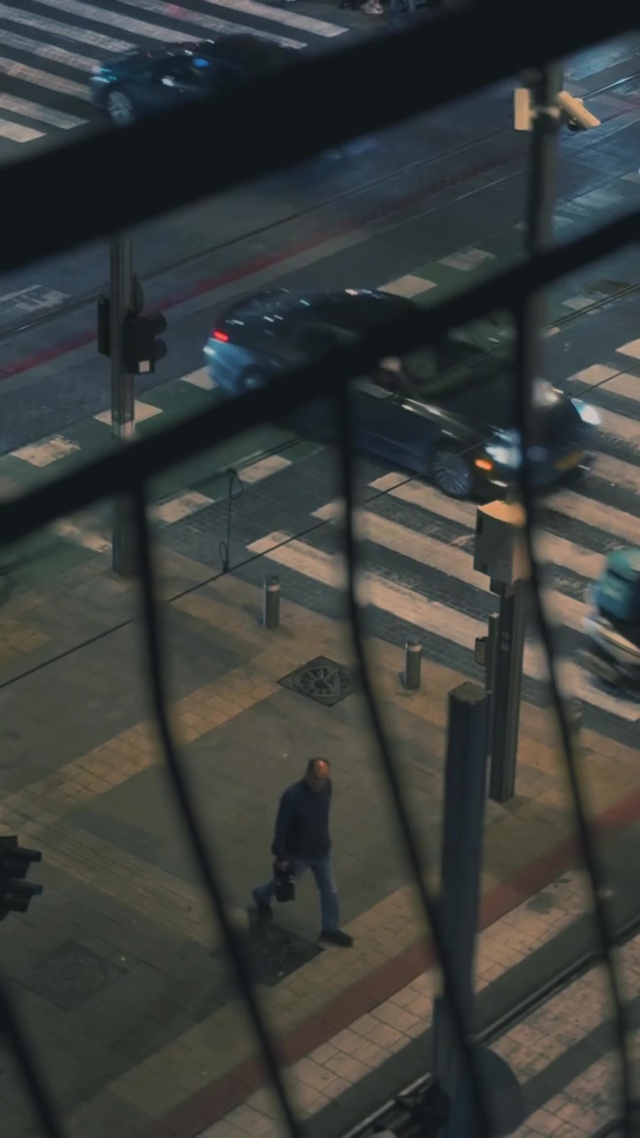 A muted city crosswalk viewed through window bars, showing sparse early morning pedestrians and passing cars in soft, warm light. Vertical video, ideal for social media.
