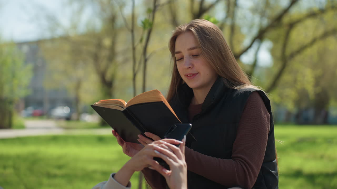 Donna caucasica che legge a un'amica su una panchina erbosa del parco, girando le pagine di un romanzo consumato, la compagna è sdraiata, luce del sole che filtra tra i giovani alberi, atmosfera primaverile calda, narrazione intima e sereno relax