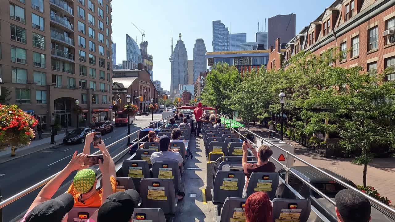 First-person onboard view of tourists exploring Toronto downtown from an open-top bus, featuring the iconic skyline, bustling streets, and a lively urban atmosphere under clear blue skies, Canada
