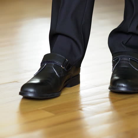 man's legs shows black shoes standing on heels and on socks in turn on the wooden floor. legs of a teenager in black trousers and the dancing shoes. close-up