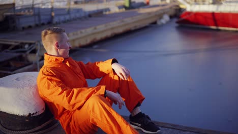 Junger-Mann-In-Orangefarbener-Uniform-Sitzt-Während-Seiner-Pause-Am-Meer-Im-Hafen