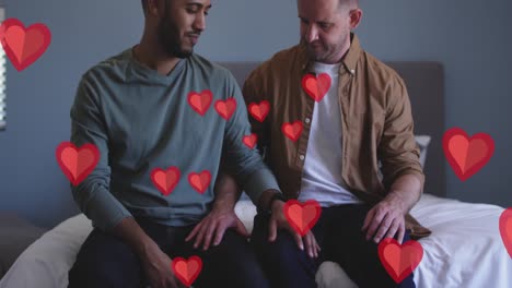 Animation-of-red-hearts-over-diverse-male-couple-sitting-on-bed