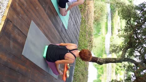 Toma-Vertical-De-Damas-Haciendo-Yoga-Meditando-Frente-A-Un-Lago-Claro-Y-Tranquilo