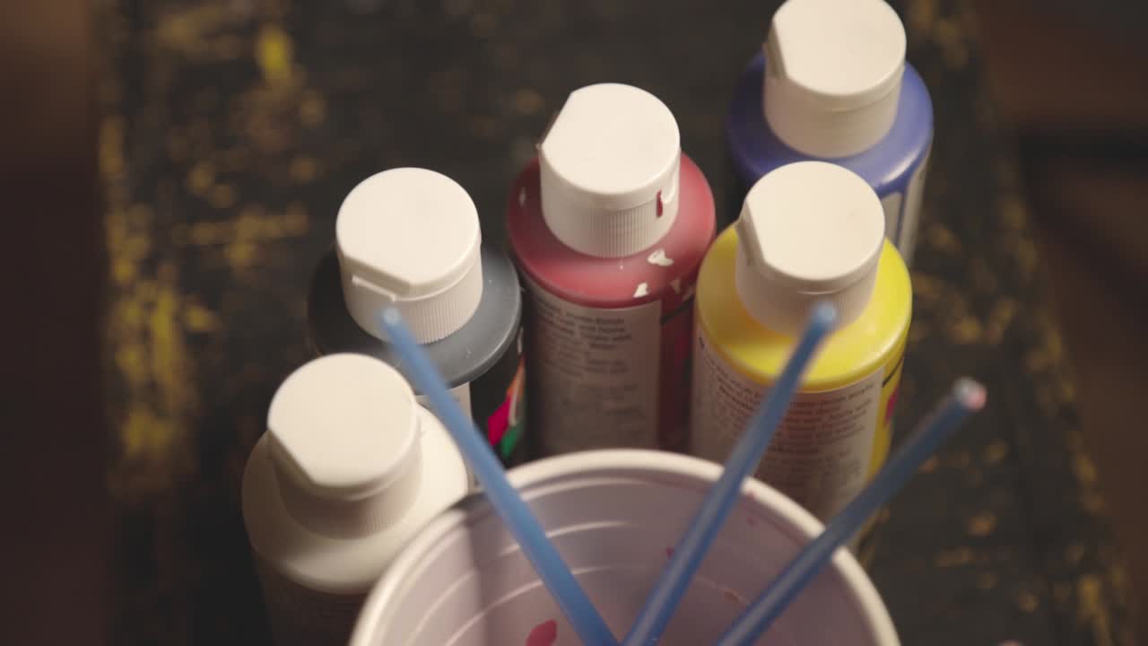 Used Paintbrushes In A Plastic Cup With Many Colorful Paint Bottles On The Wooden Table.- closeup shot