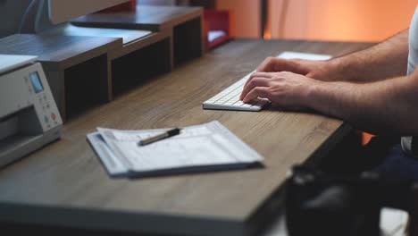 creative business person sitting at his home office modern wood desk typing on a white wireless computer keyboard, clicking a mouse trackpad next to a printer, monitor and camera and then walks away