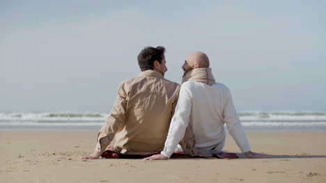 Vista-Trasera-De-Una-Alegre-Pareja-Homosexual-Inclinando-La-Cabeza-El-Uno-Al-Otro-Mientras-Se-Sientan-En-La-Playa