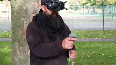 hombre barbudo con gafas fpv de realidad aumentada que controla drones en el parque con joystick de movimiento