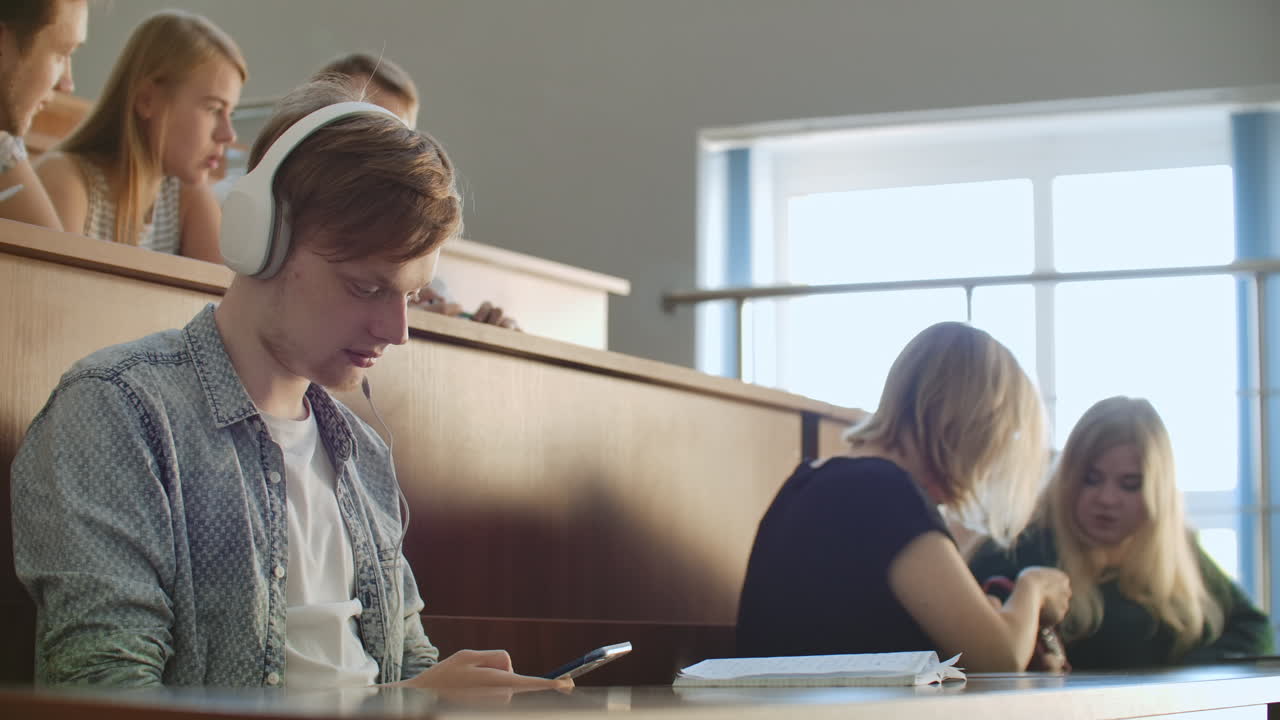 A male student in a University auditorium listens to music in white headphones during a break without paying attention to others. A lot of people in the audience are talking.